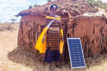Eine Frau in traditioneller Kleidung steht vor einer Lehmhütte und einem Solarpanel in einer trockenen Landschaft. Sie lächelt und trägt ein gelbes Tuch über ihrer Schulter.