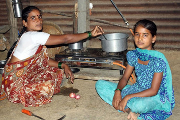 Une femme et une jeune fille sont assises par terre dans une cuisine simple. La femme cuisine sur un réchaud à gaz, tandis que la fille est assise à côté d'elle. Deux oignons et un couteau sont posés sur le sol.