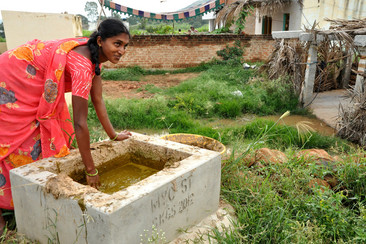 Une femme en sari rouge se penche sur un réservoir rectangulaire en ciment rempli d'eau. En arrière-plan, on voit des champs verts et une maison simple.