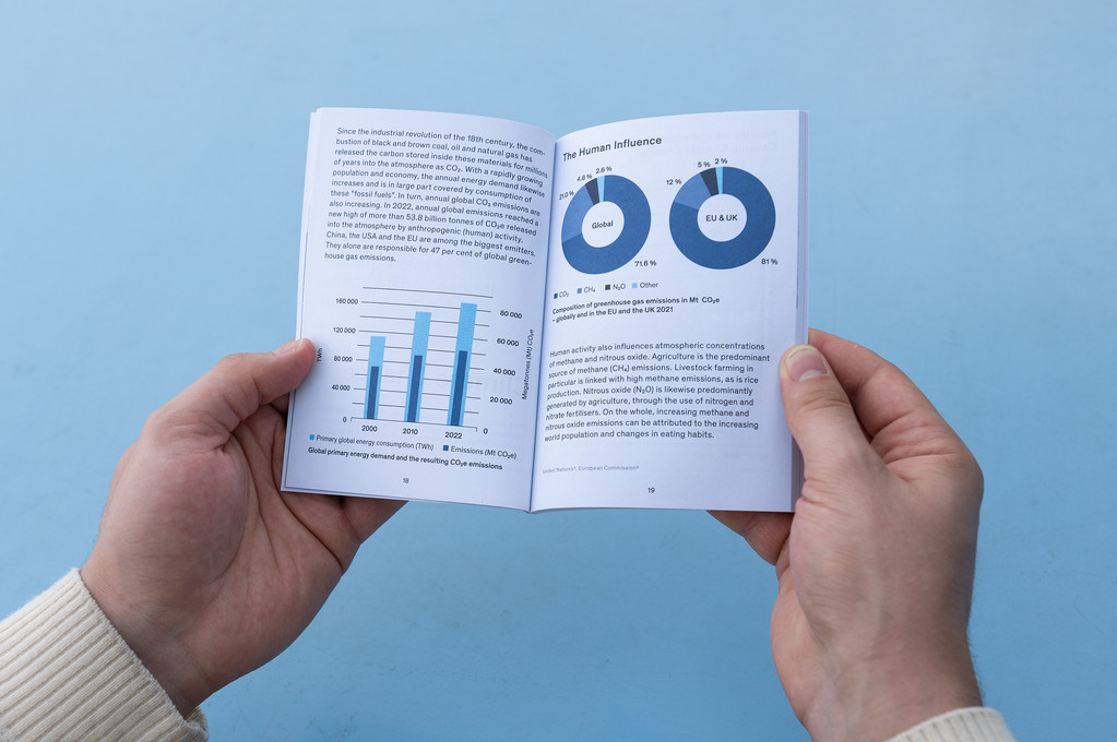 Zwei Hände halten ein offenes Faltblatt, das Diagramme und Statistiken in Blau zeigt. Der Hintergrund ist einfarbig hellblau.