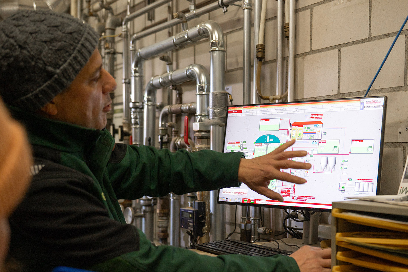 A man in a cap points to a monitor with a control system for a biogas digester. In the background, numerous stainless steel pipes run through the plant room.