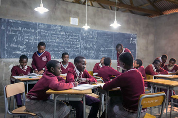 Un groupe d'élèves en uniformes scolaires bordeaux est assis dans une salle de classe et étudie ensemble. En arrière-plan, on voit un tableau noir avec des notes et des formules écrites, et plusieurs lampes pendent du plafond.