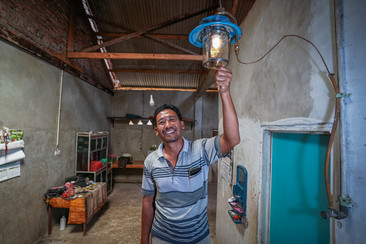 Un uomo sorride e si trova in una stanza arredata in modo modesto con pareti rustiche di mattoni e intonaco. Tiene in mano una lampada blu accesa che pende dal soffitto e proietta una luce calda sull'ambiente circostante. Sullo sfondo c'è una semplice porta di legno e uno scaffale ben organizzato con oggetti di uso quotidiano.