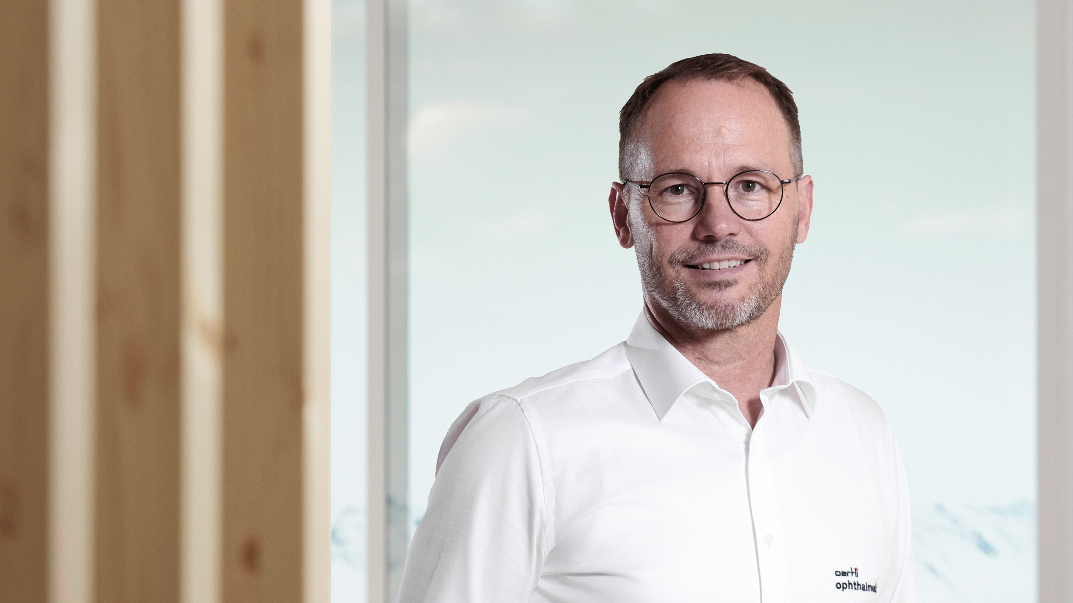 Man wearing glasses and a white shirt stands in front of a window with the words ‘Eye Surgery Essentials’ written on it; snow-covered Alps and wooden structures are visible in the background.