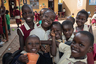 A group of smiling children in school uniforms joyfully posing for the camera in a lively schoolyard.