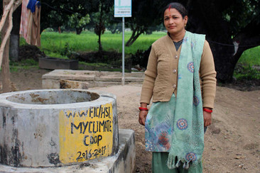 A woman stands next to a masonry well in a rural setting, with inscriptions visible on the well.