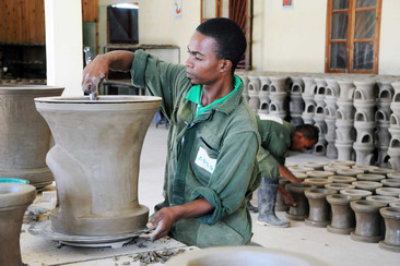 Un homme en tenue de travail verte façonne un grand pot en céramique dans un atelier. En arrière-plan, on voit un autre ouvrier travaillant avec plusieurs pots.
