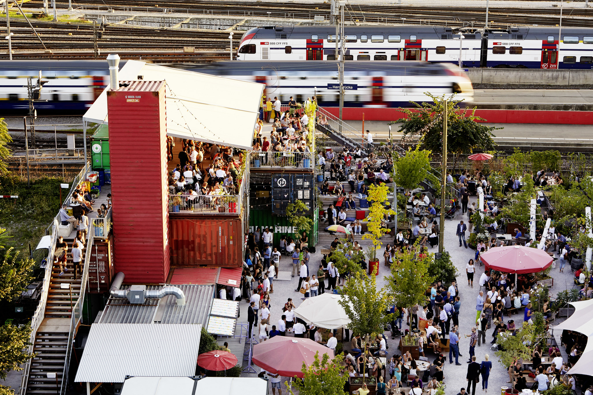 Vue aérienne d’un événement en plein air animé près des voies ferrées. Des personnes se rassemblent sur une place avec des parasols et des arbres, un train passe à l’arrière-plan. Un bâtiment rouge avec une terrasse se trouve à gauche.