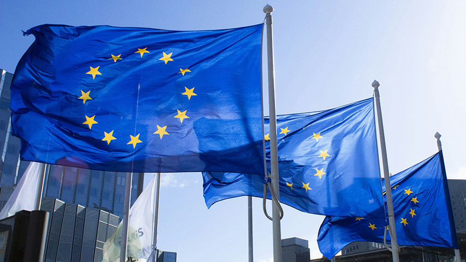 Drei wehende EU-Flaggen mit gelben Sternen auf blauem Hintergrund vor modernen Bürogebäuden an einem sonnigen Tag in Brüssel.