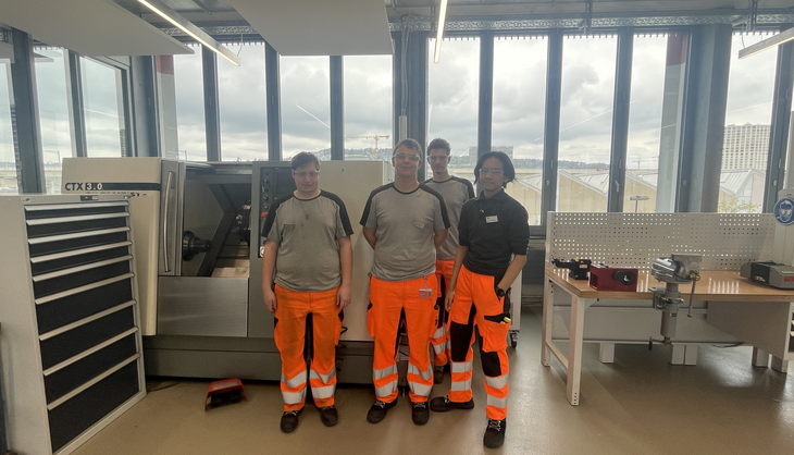 Vier Arbeiter in orangenen Warnschutzhosen stehen in einem modernen Werkraum mit grossen Fenstern und Industriemaschinen. Sie tragen graue und schwarze Arbeitskleidung und blicken in die Kamera.