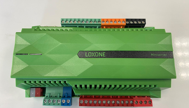 Grünes Loxone-Steuergerät mit mehreren farbigen Anschlüssen, auf einem weissen Hintergrund liegend. Das Gerät ist für Gebäudeautomation konzipiert.