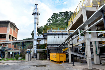 Eine industrielle Anlage mit einem hohen Schornstein, gelben Containern und mehreren Gebäuden. Im Hintergrund sind Bäume und Hügel zu sehen.