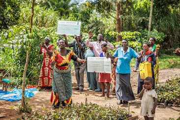 A group of people stands in a garden holding signs while smiling cheerfully at the camera. A small child stands in the foreground, looking to the side.