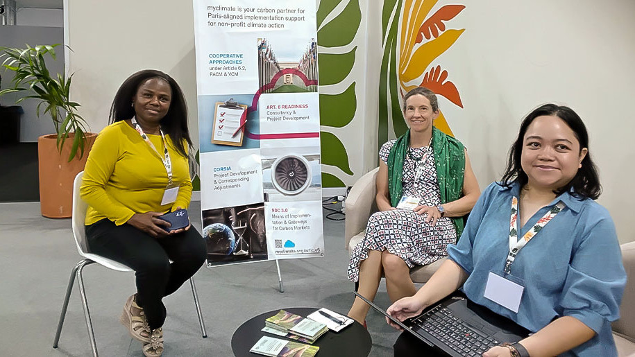 Drei Frauen sitzen in einem klimafreundlich gestalteten Messestand mit Pflanzenmotiven und Informationsplakaten zum CO₂-Markt. Eine Frau hält ein Laptop, auf dem Tisch liegen Flyer zur Klimafinanzierung. Im Hintergrund steht ein Roll-up mit Texten zu internationalen Klimaschutzmechanismen.