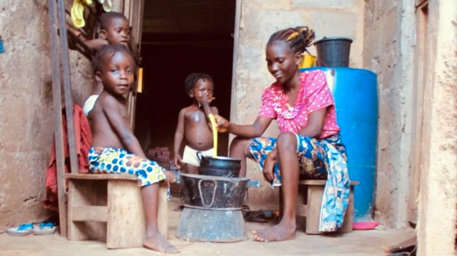 A woman and three children sit next to a cooking stove and in front of a house.