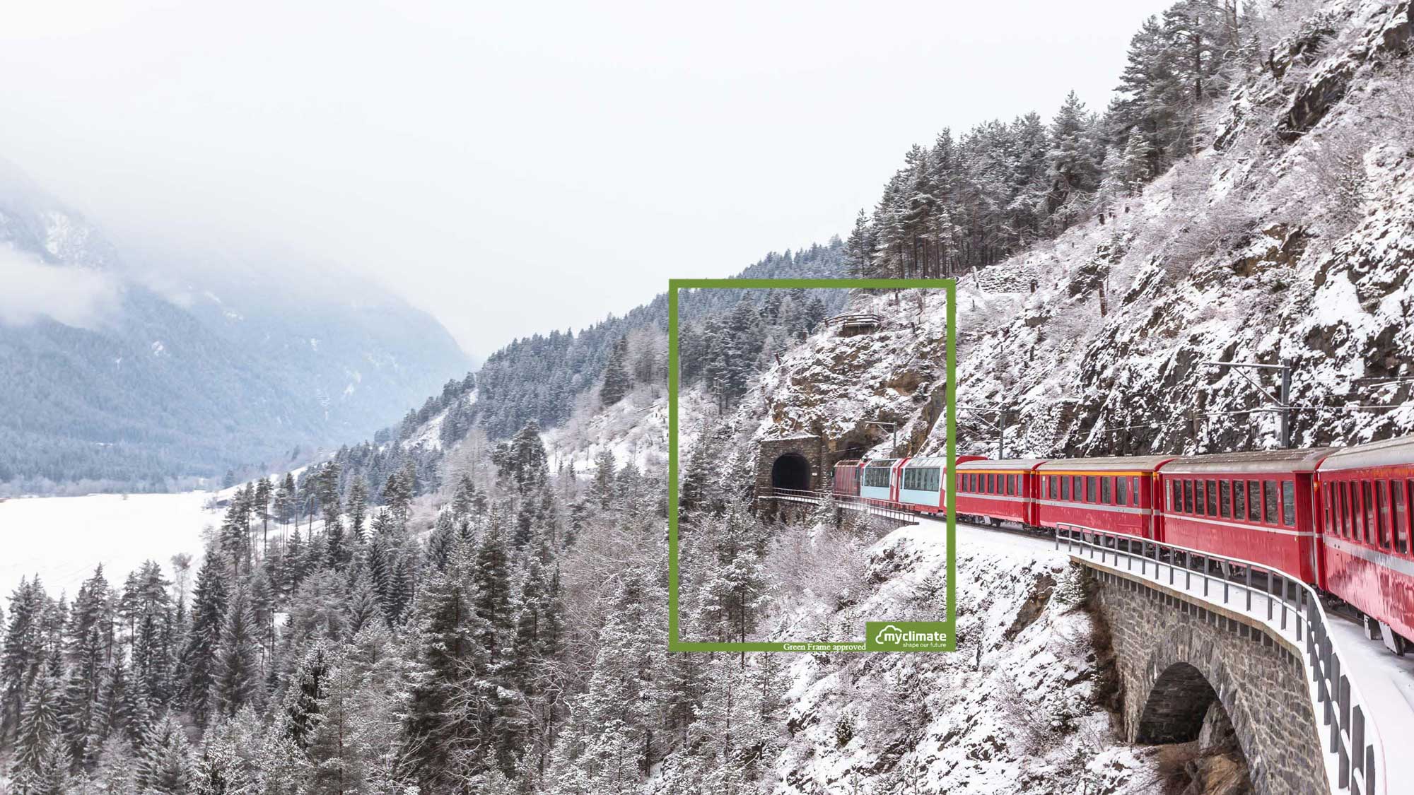 Ein roter Zug fährt durch eine schneebedeckte Landschaft in den Schweizer Alpen. Der Zug bewegt sich entlang eines Steinviadukts und nähert sich einem Tunnel in einem bewaldeten Berghang. Umgeben von verschneiten Tannen, bietet das Bild eine winterliche, ruhige Atmosphäre mit nebelverhangenen Bergen im Hintergrund.