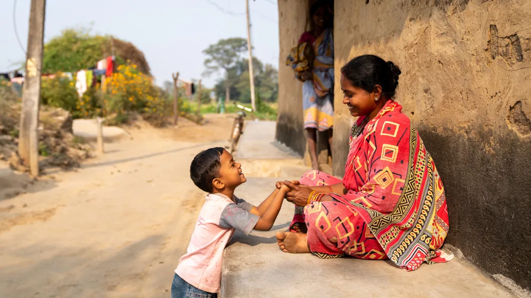 A woman in a colourful sari sits barefoot on a step in front of a mud house and lovingly holds the hands of a laughing little boy. In the background, another person stands in front of the wall of the house, and a rural road leads into the countryside.