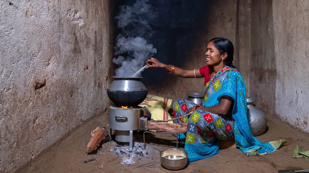 A woman in a colourful sari sits on the floor of a simple kitchen with unplastered walls and cooks over a smoking wood-burning stove. She smiles as she stirs a large pot, while metal bowls and kitchen utensils stand around her.