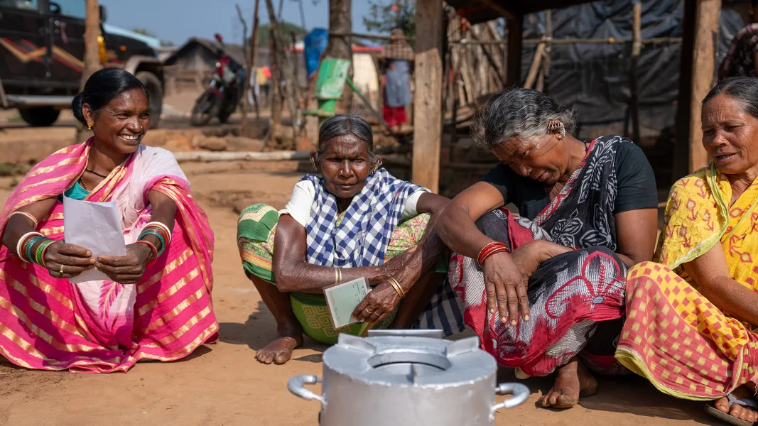 Four women in brightly patterned saris sit outside on the ground in a village, some with documents in their hands. In front of them is a modern metal cooker. Huts, a motorbike and a car are visible in the background.