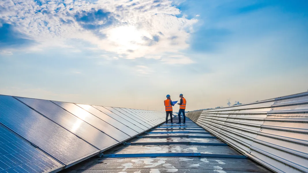 Zwei Techniker in Warnwesten stehen auf einem Dach mit grossflächigen Solarpaneelen. Sie besprechen sich unter einem bewölkten Himmel, während die Sonne auf den Paneelen reflektiert wird.
