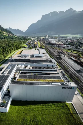 Luftaufnahme eines Industriegebäudes mit Photovoltaikanlagen auf dem Dach neben Bahntrassen und einer Stadtlandschaft mit Bergkulisse im Hintergrund.
