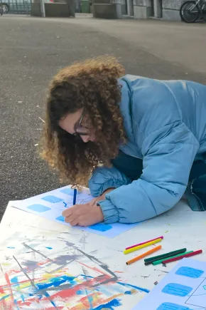 Eine Person sitzt im Freien am Boden und arbeitet kreativ mit Papier und Stiften.