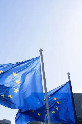 Zwei Europaflaggen, blau mit Sternen wehen vor hellblauem Himmel.