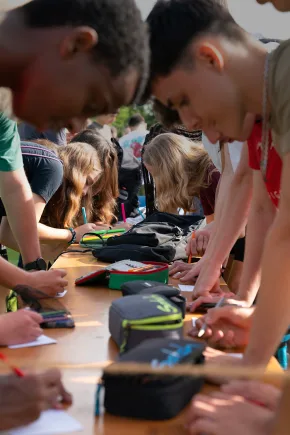 Mehrere junge Menschen stehen im Freien an einem Tisch und schreiben oder zeichnen gemeinsam auf Papier.