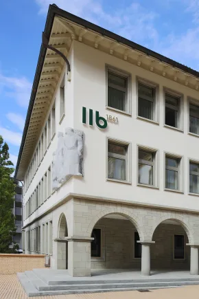 Ein schickes, älteres Gebäude mit Säulen im Erdgeschoss und Logo auf der Fassade vor einem blauen Himmel.